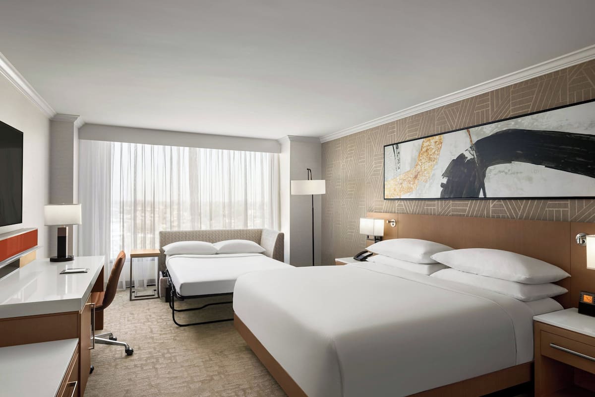 A hotel room with two beds, a desk, a chair, a TV, and a large abstract painting on the wall.