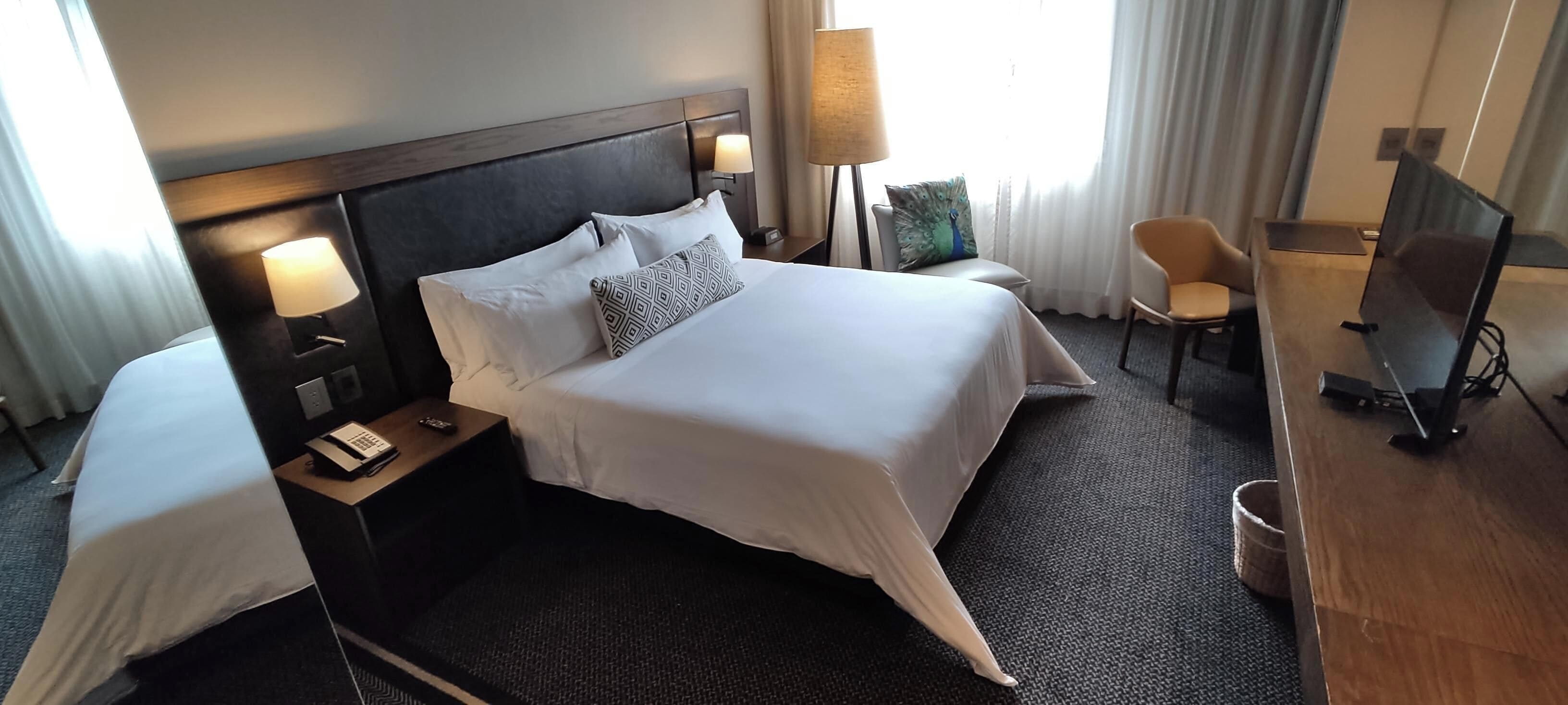 Ein Hotelzimmer mit einem Bett, Nachttischlampen, einem Schreibtisch mit Computer, einem Sessel und einem Fernseher.