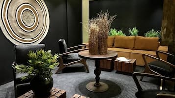 A modern lounge area with a round table, a sofa, and chairs, featuring a decorative wall piece and potted plants.