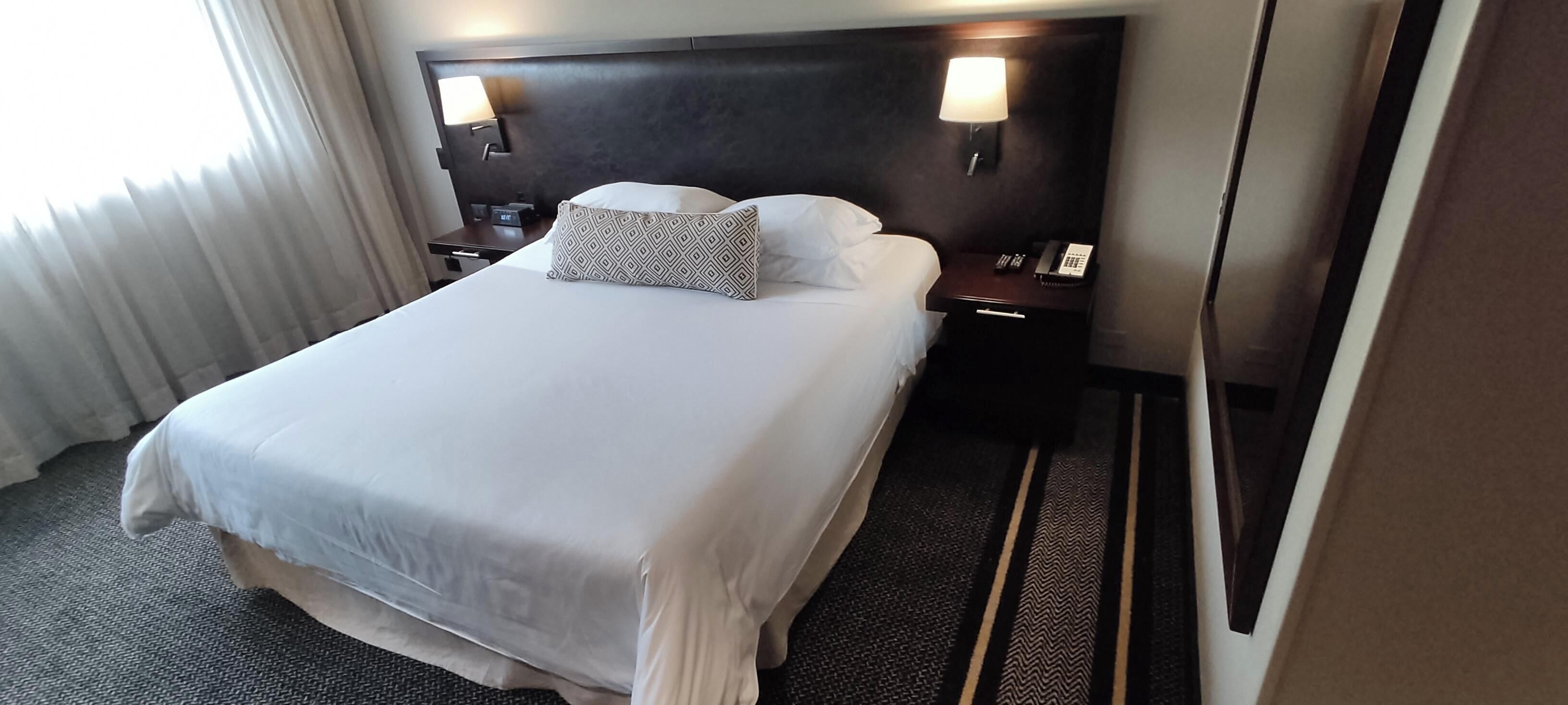 A hotel room with a large bed, two bedside tables with lamps, a nightstand with a phone, and a window with curtains.