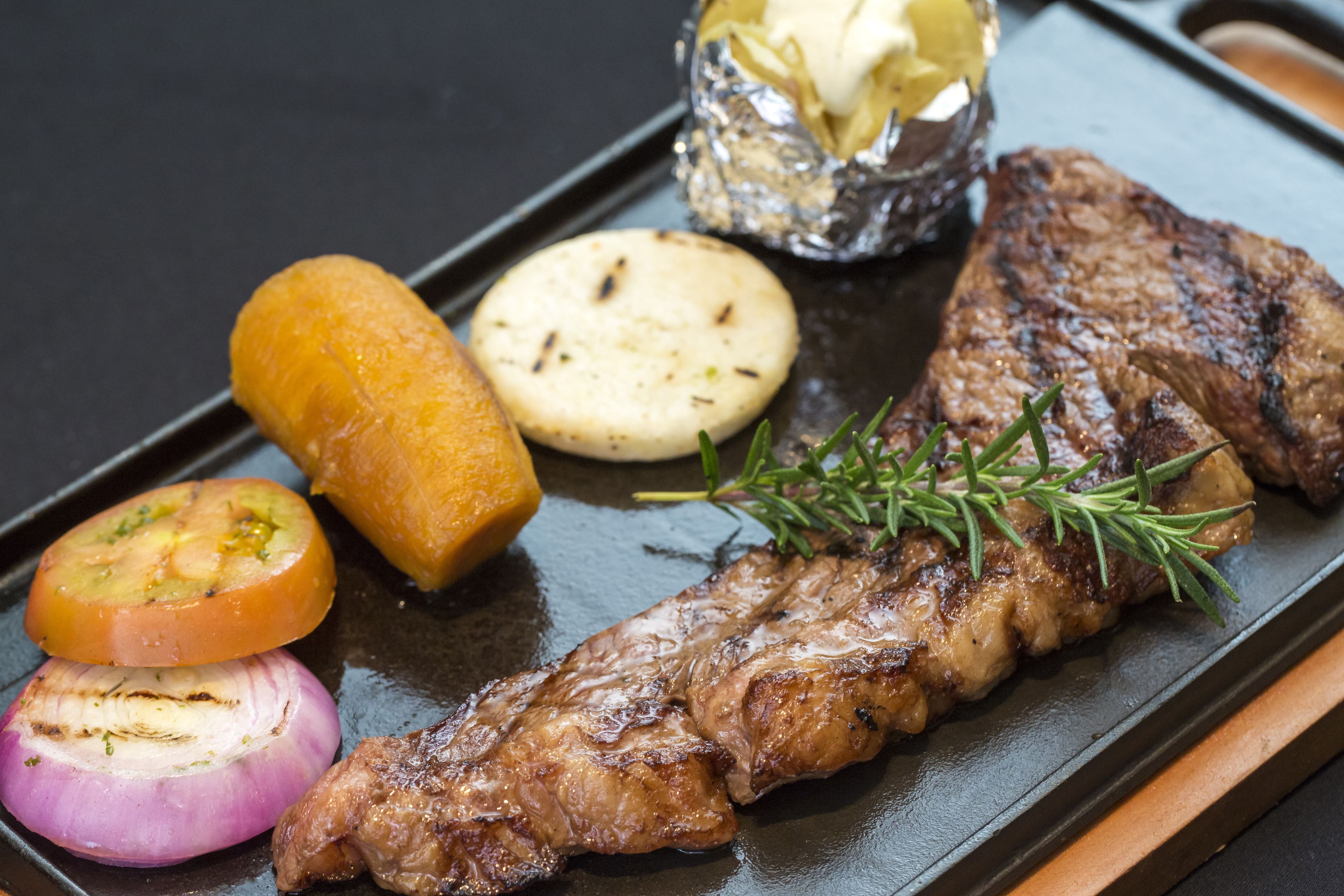 Un filete acompañado de verduras a la parrilla y un recipiente pequeño con mantequilla sobre un plato negro.