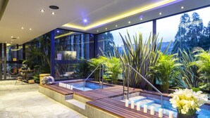 A modern indoor pool area with wooden decking, a hot tub, and a view of the surrounding greenery.