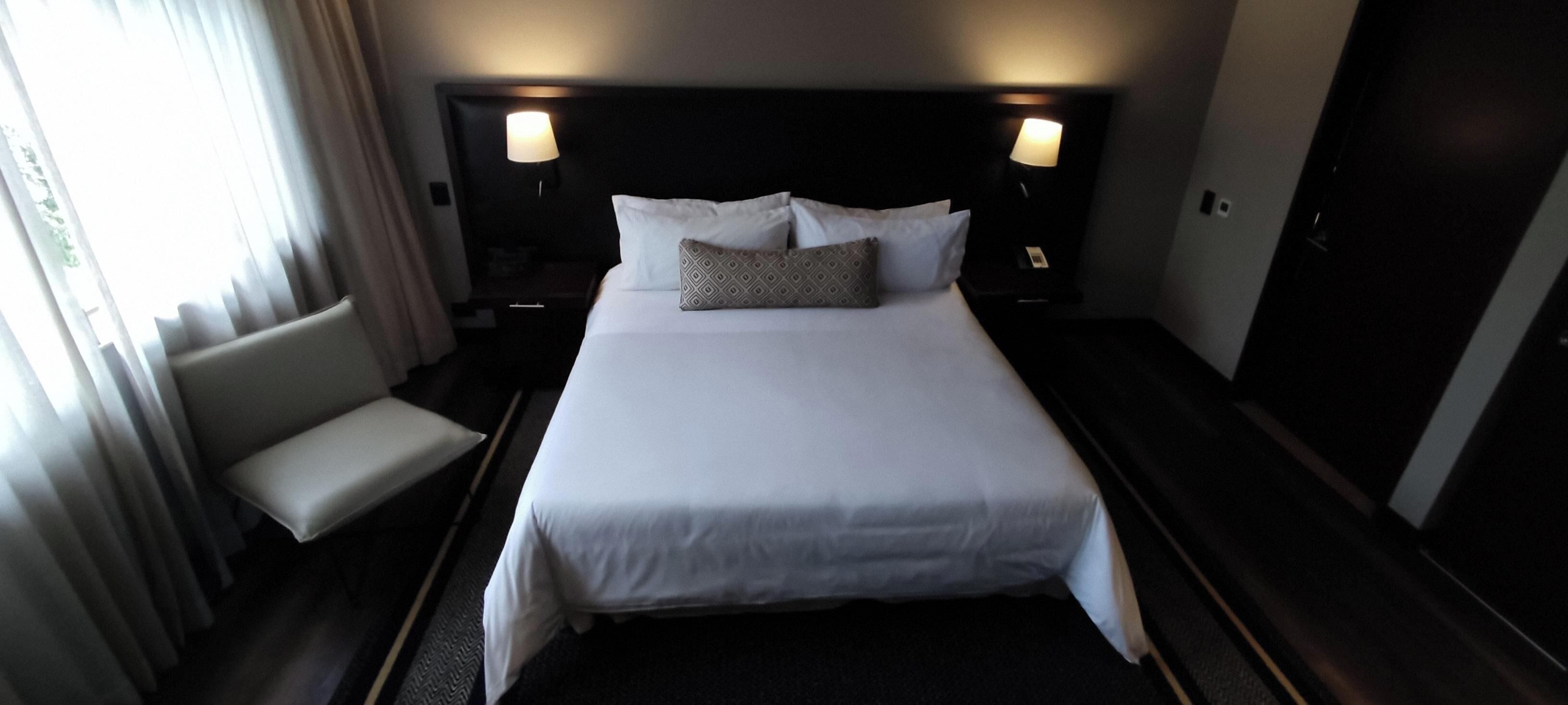 A hotel room with a large bed, bedside lamps, a bench, and a wardrobe.