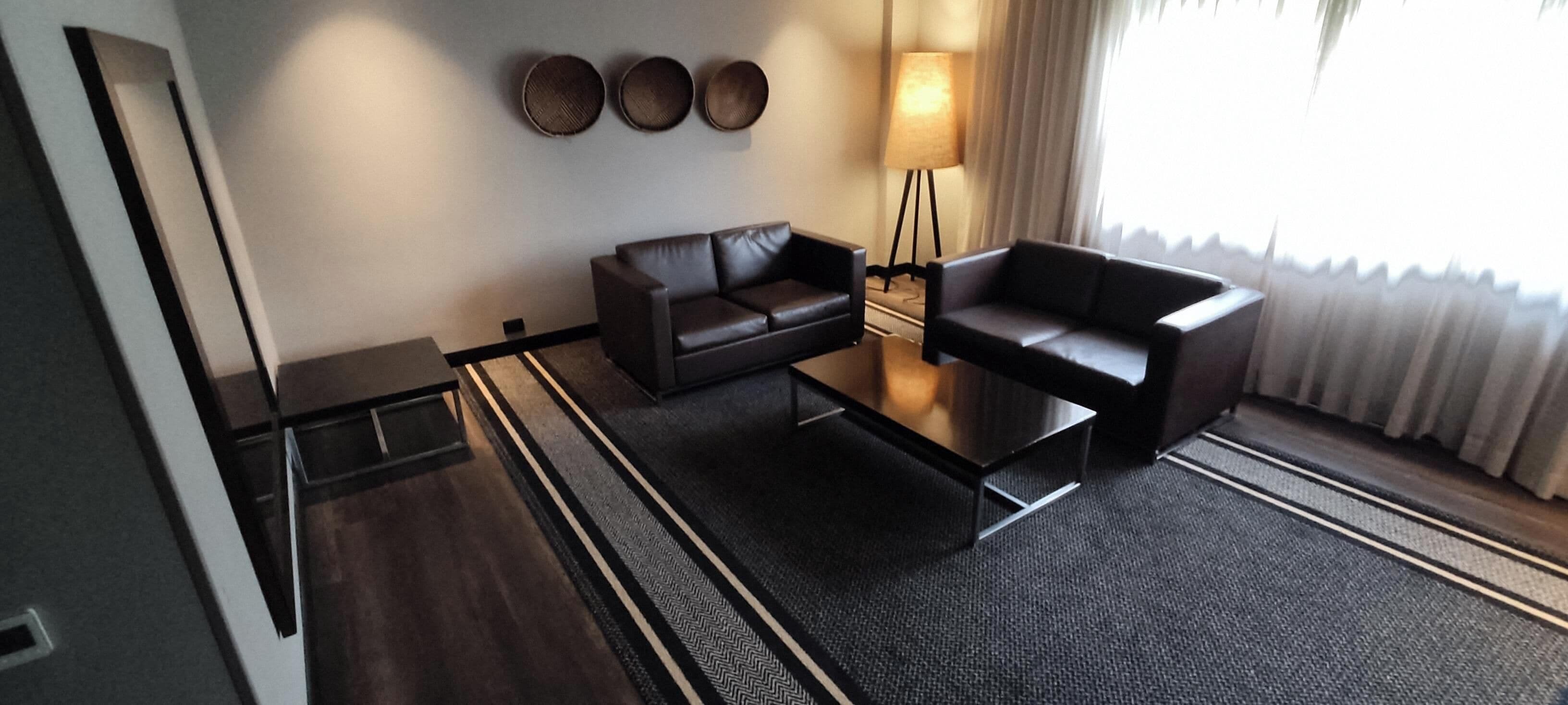 Une chambre d’hôtel moderne avec un canapé en cuir foncé, un fauteuil assorti, une petite table basse et un lampadaire. Trois assiettes décoratives sont accrochées au mur au-dessus du canapé.