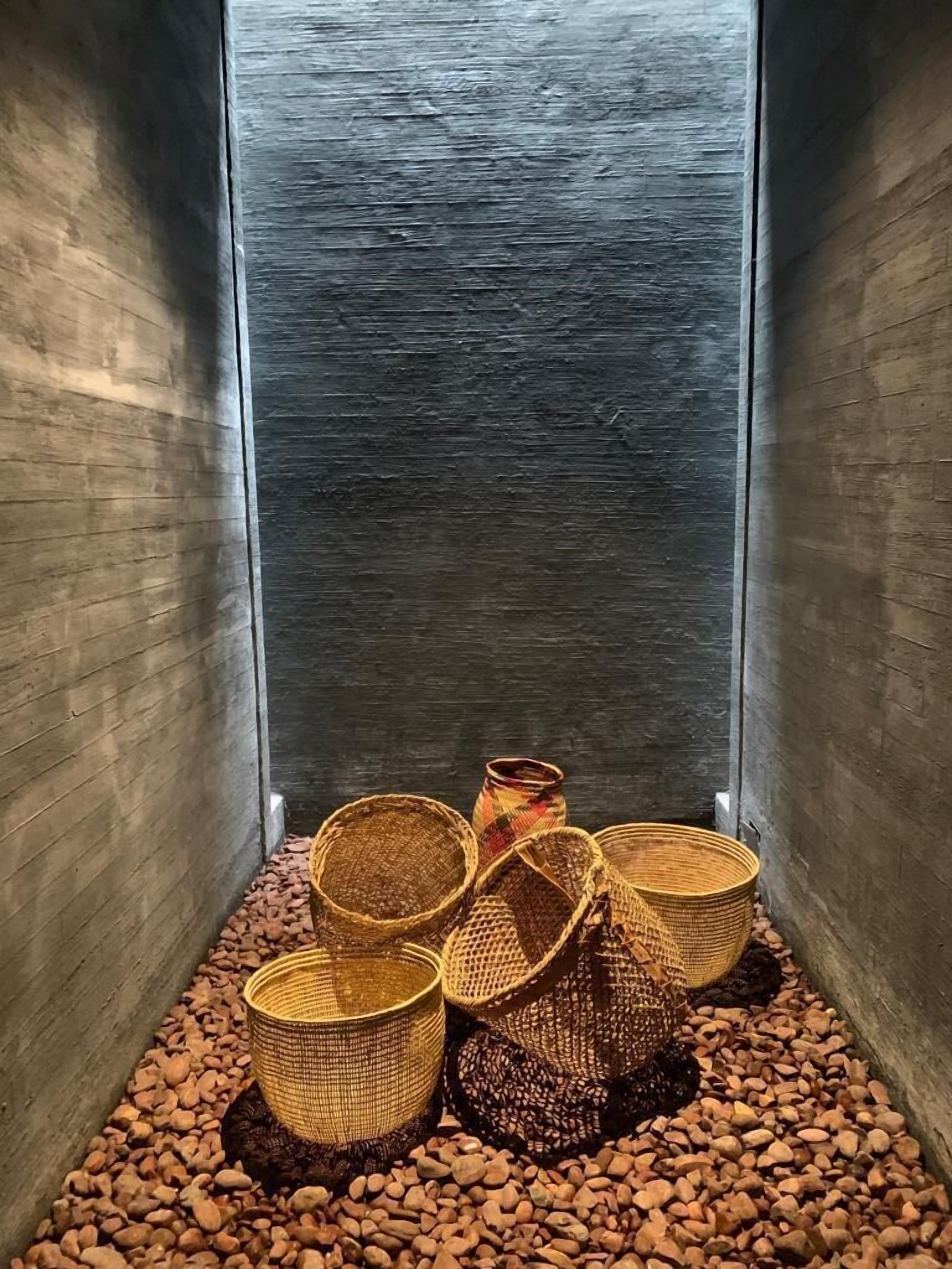 Un piccolo ambiente raccolto con un muro strutturato, diverse ceste intrecciate su un pavimento di ciottoli e una piccola pianta colorata in vaso.