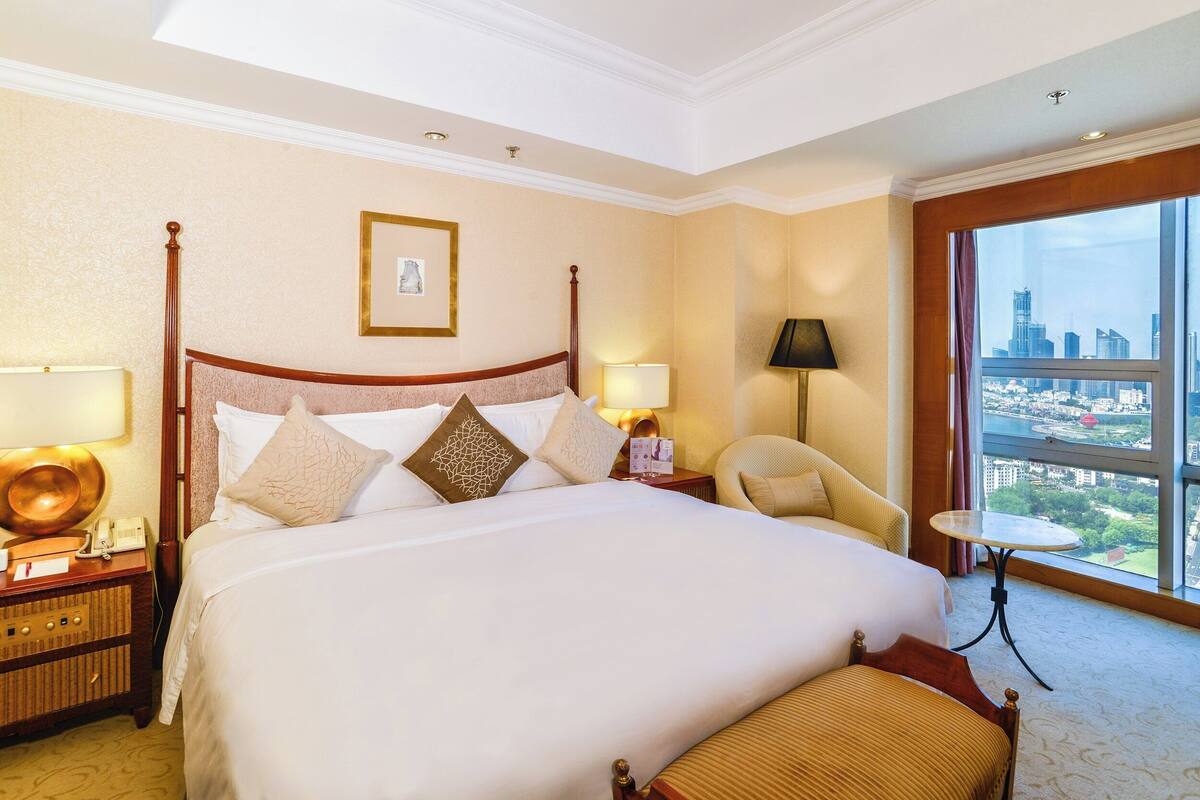 Een hotelkamer met een groot bed, een nachtkastje met lamp, een stoel, een klein tafeltje en uitzicht op een stadsbeeld door een groot raam.