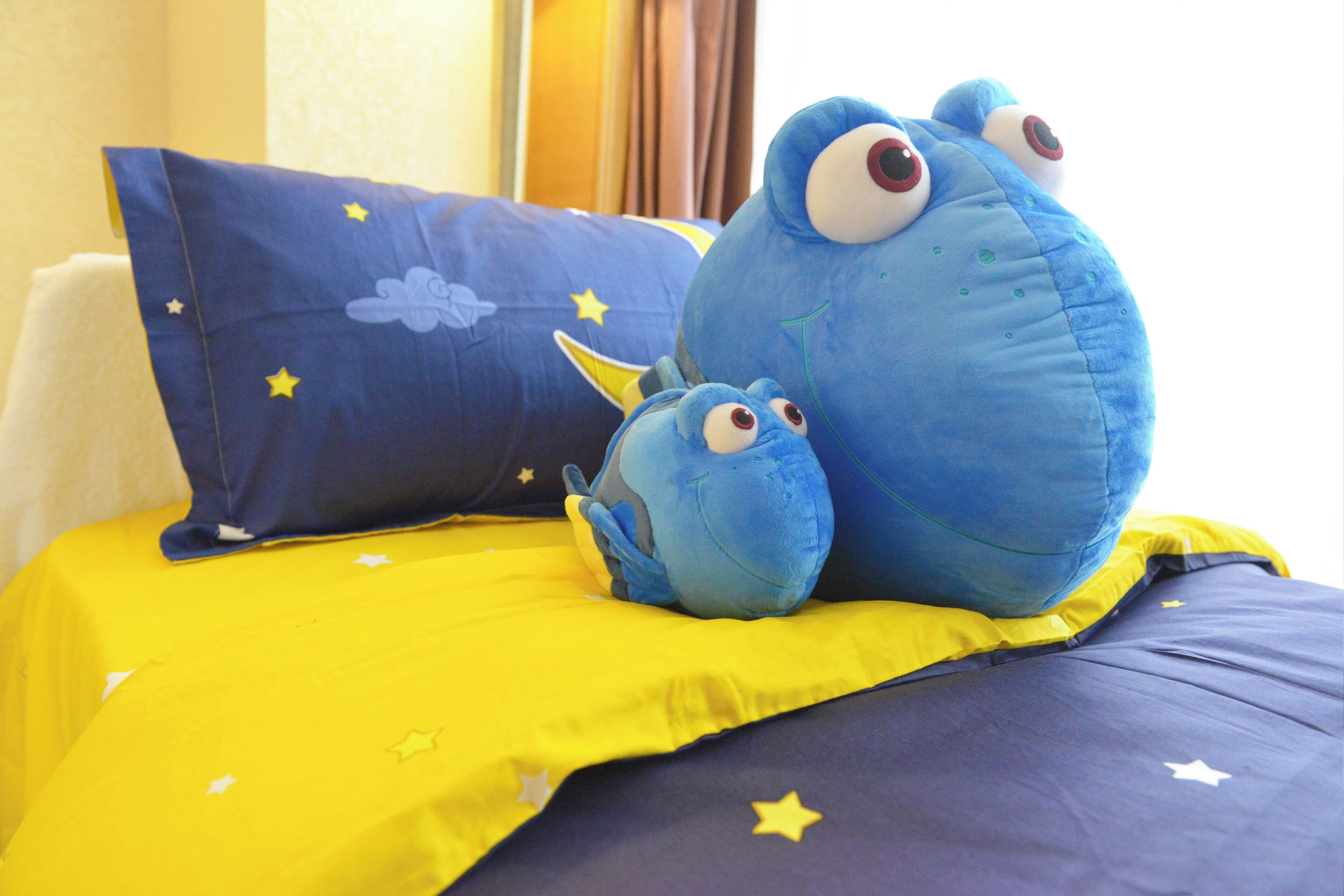 A bed with a yellow and blue bedspread, two blue plush toys, and a blue pillow with stars.