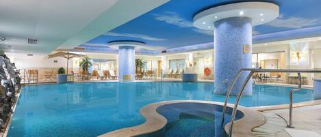 Piscina interior con diseño de mosaico azul, sillas de descanso y techo con mural que simula el cielo.