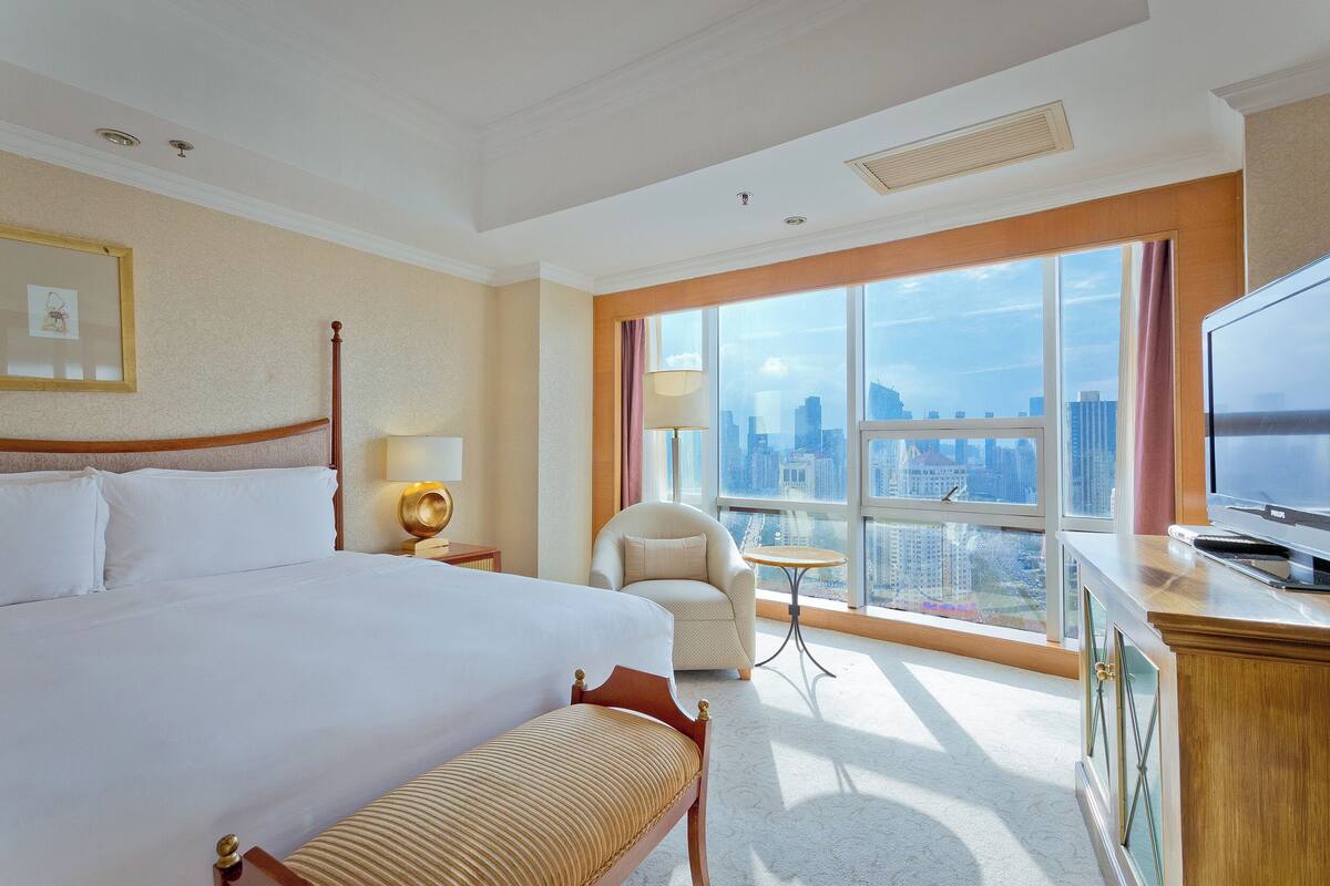 Hotelkamer met een groot bed, een stoel, een bureau met een televisie en een groot raam met uitzicht op de stad.