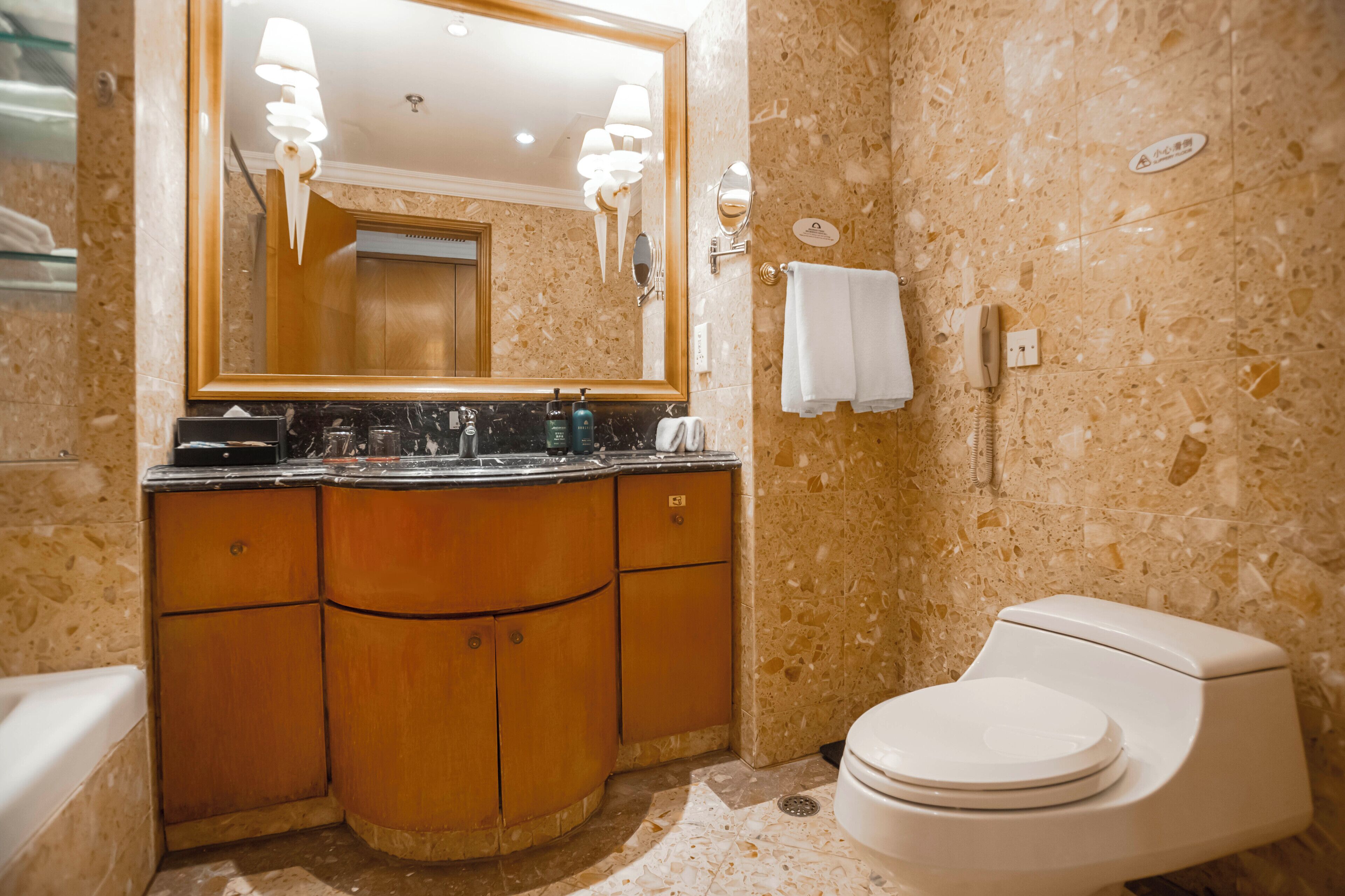 A bathroom with a marble wall, a wooden vanity, a toilet, and a mirror with lights.