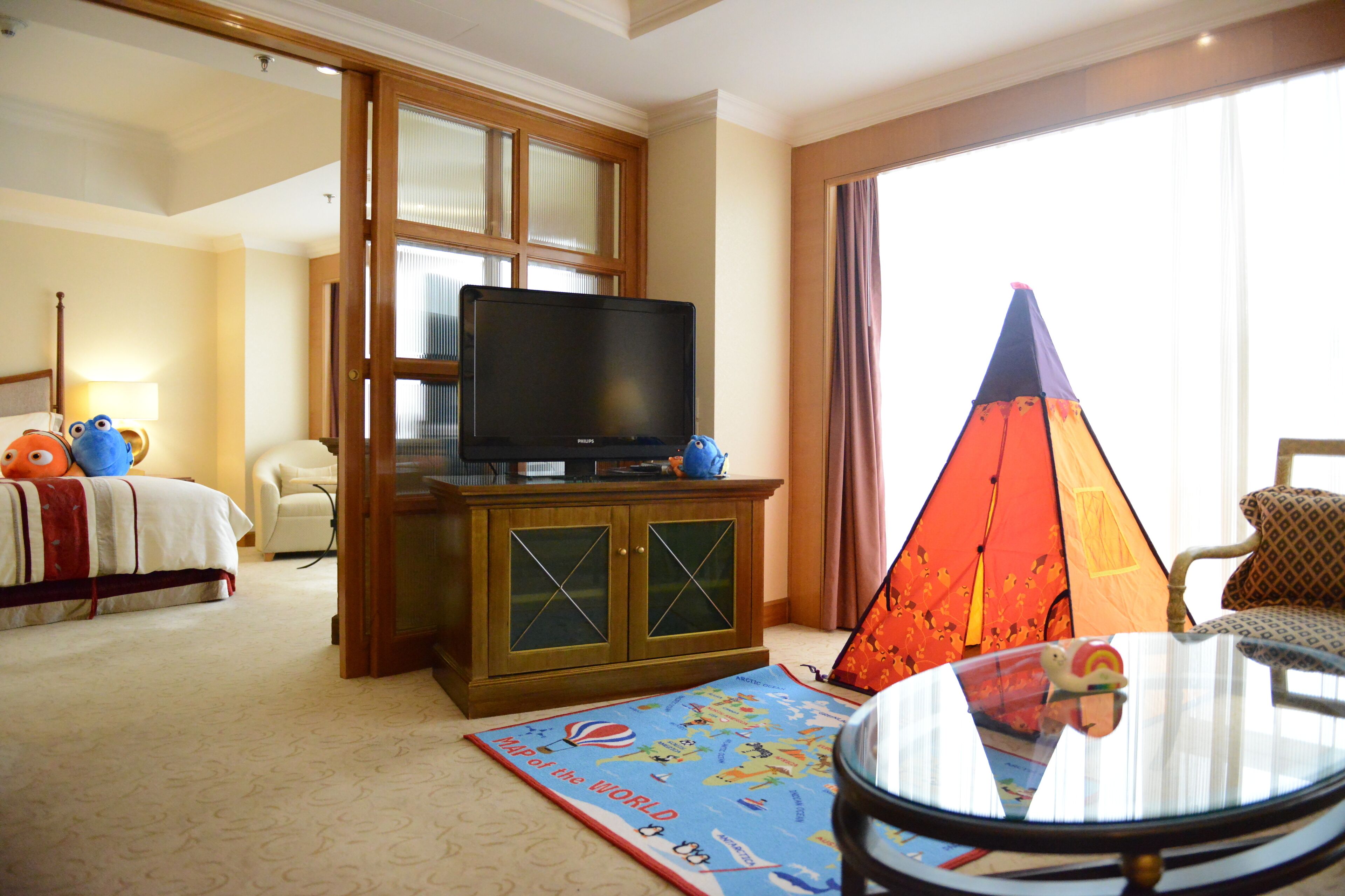 Quarto de hotel com uma cama, uma televisão sobre um móvel de madeira, um tapete com um mapa, uma tenda e uma mesa redonda com um brinquedo.