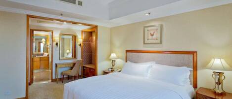 A hotel room with a large bed, a desk with a chair, a mirror, a bedside table with a lamp, and a framed picture on the wall.