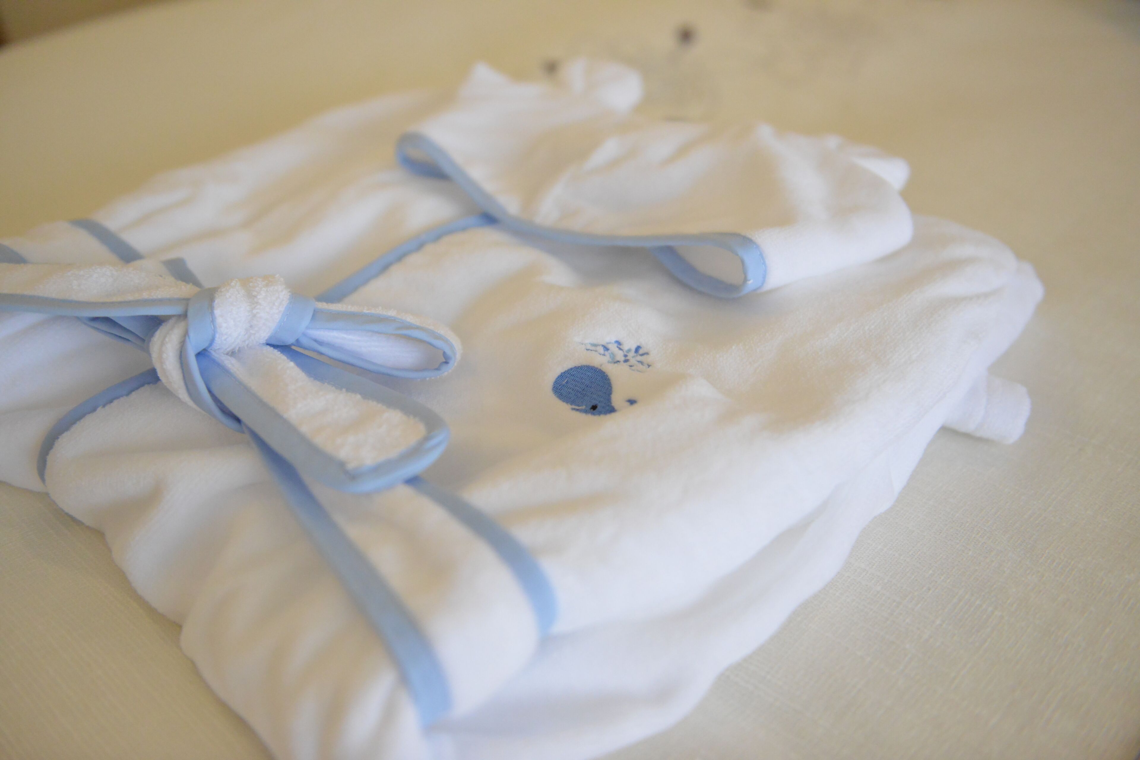 A neatly folded white towel with a blue ribbon and a small embroidered design, placed on a white surface.