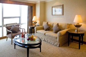 A hotel room with a sofa, armchair, two side tables with lamps, a glass coffee table with fruit, and a large window with curtains.
