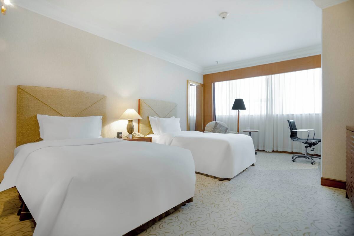 Een hotelkamer met twee bedden, een bureau, een stoel en een groot raam met gordijnen.