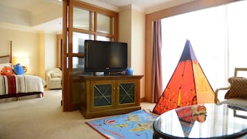 Quarto de hotel com televisão sobre um armário de madeira, cama com edredom branco, tapete com mapa do mundo, uma tenda e uma mesa redonda com tampo de vidro.