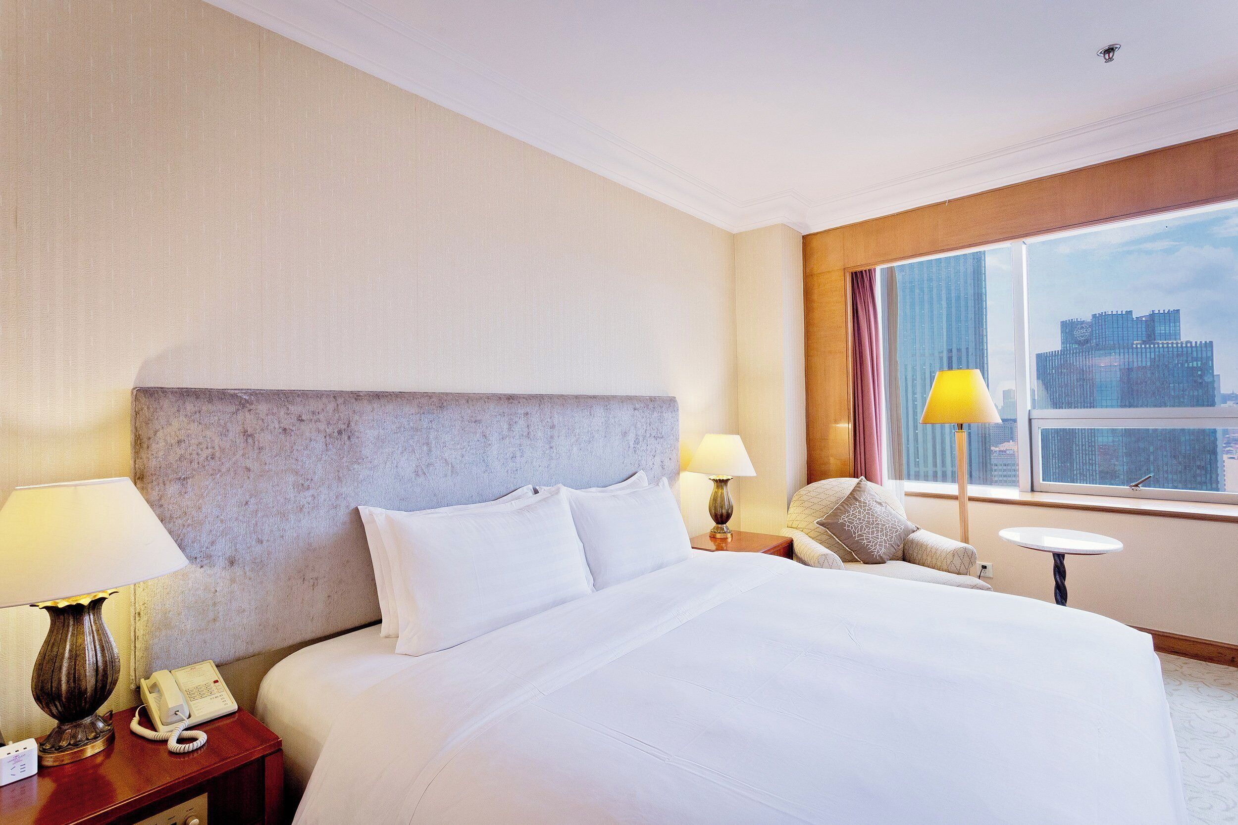 Habitación de hotel con una cama grande, una mesita de noche con lámpara, teléfono y vistas a un paisaje urbano a través de un ventanal amplio.