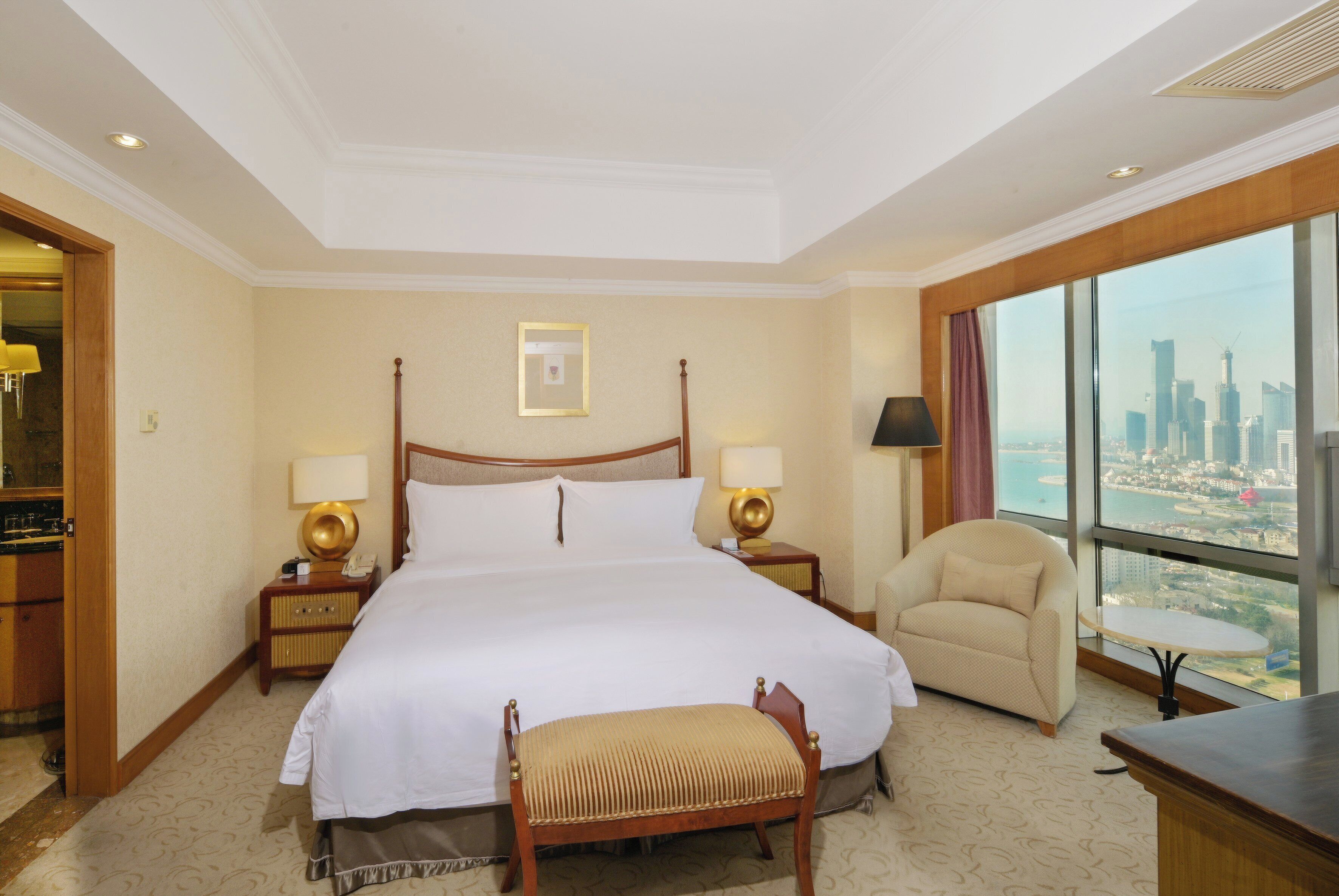 A hotel room with a large bed, two bedside tables with lamps, a chair, a small table, and a view of a cityscape through large windows.