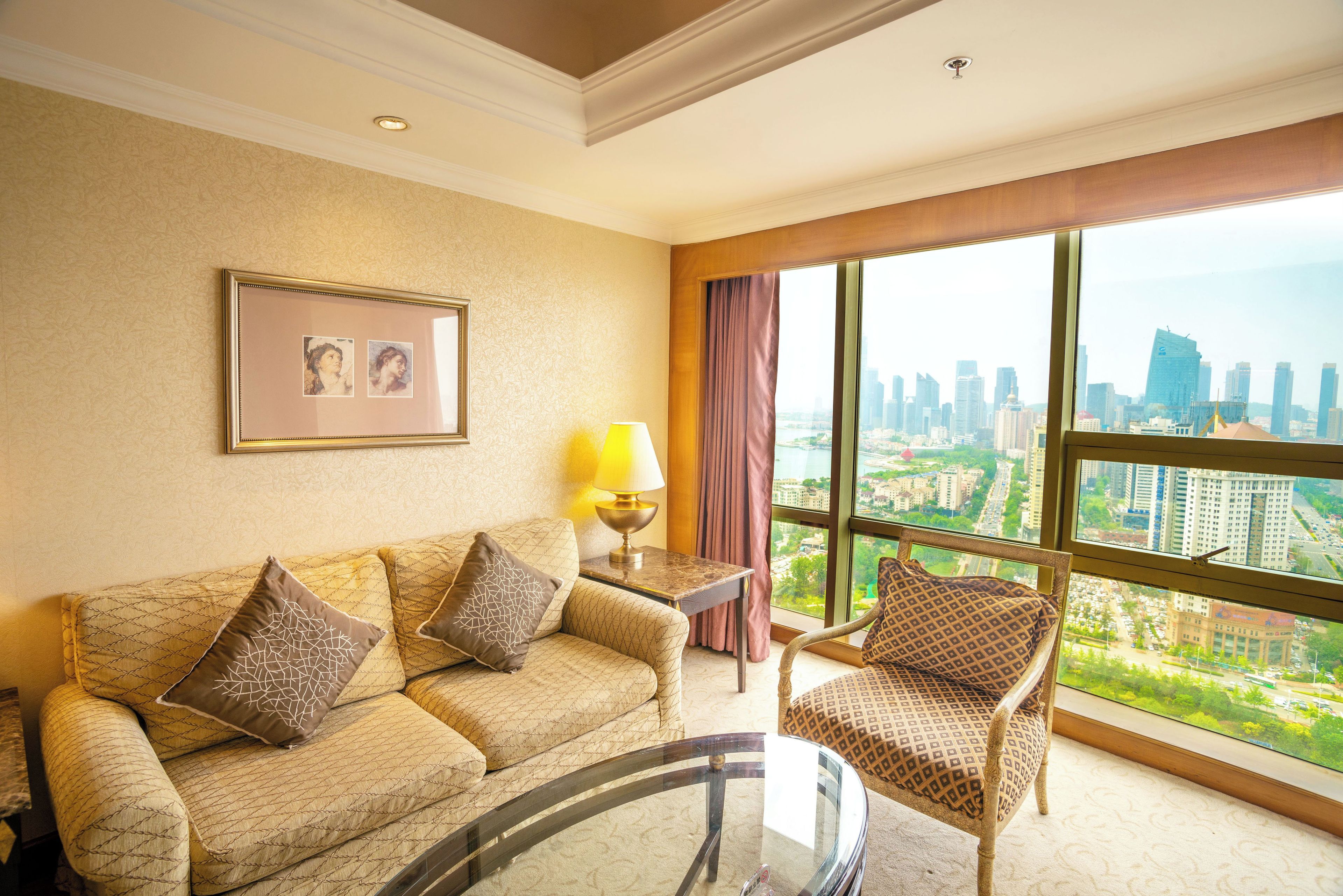A hotel room with a sofa, armchair, and a glass coffee table. There is a framed picture on the wall and a lamp on a side table.