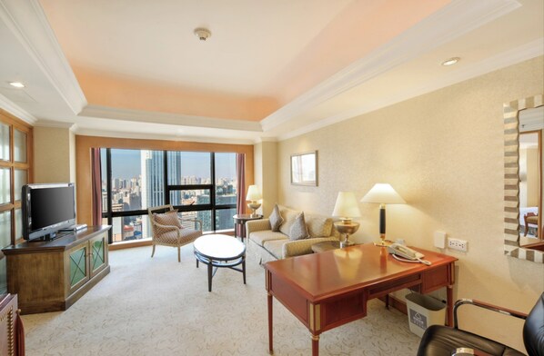 A hotel room with a city view, a television, a sofa, a coffee table, and a desk with a lamp and phone.
