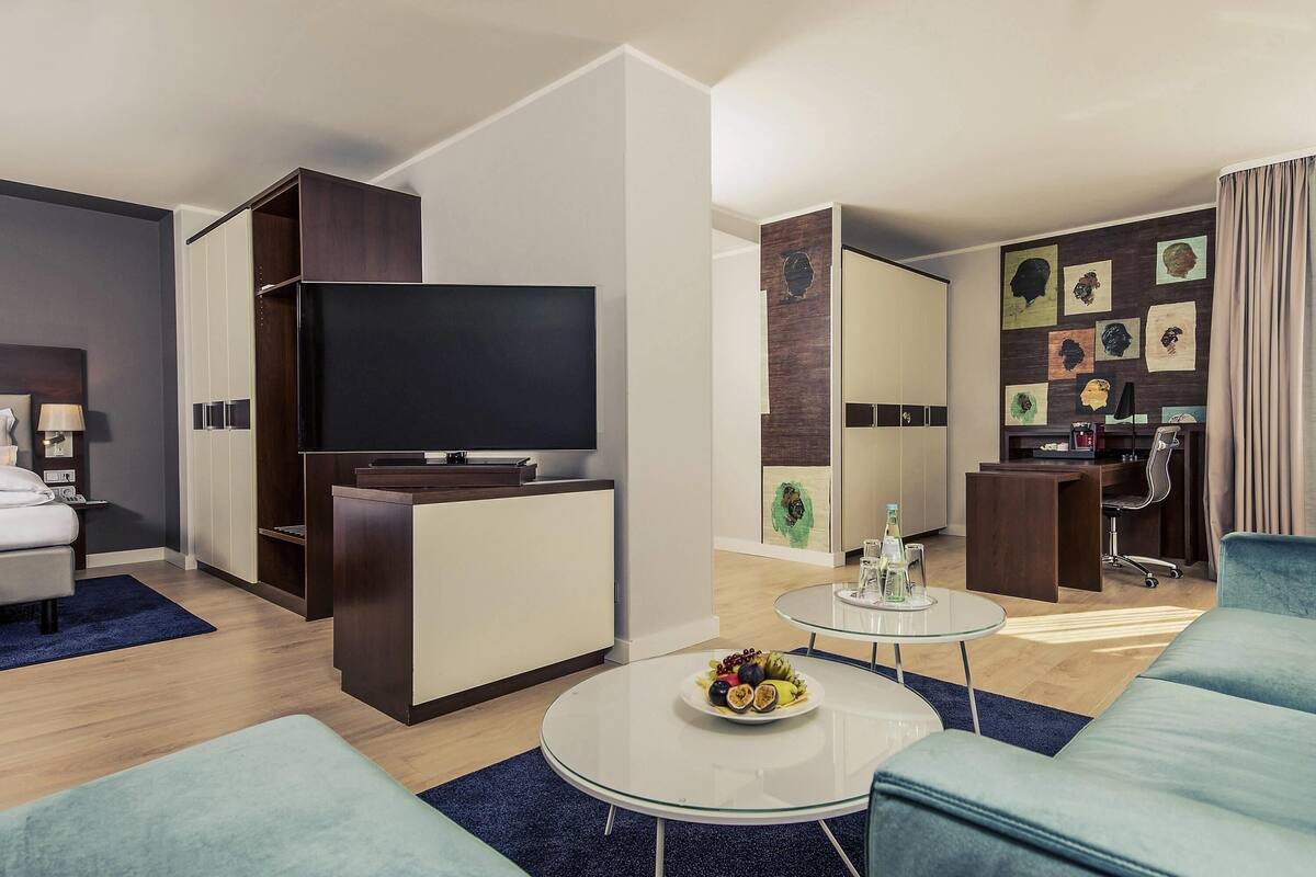 Una habitación de hotel moderna con una televisión de pantalla plana, un escritorio con silla, una mesa de comedor con fruta y un sofá.
