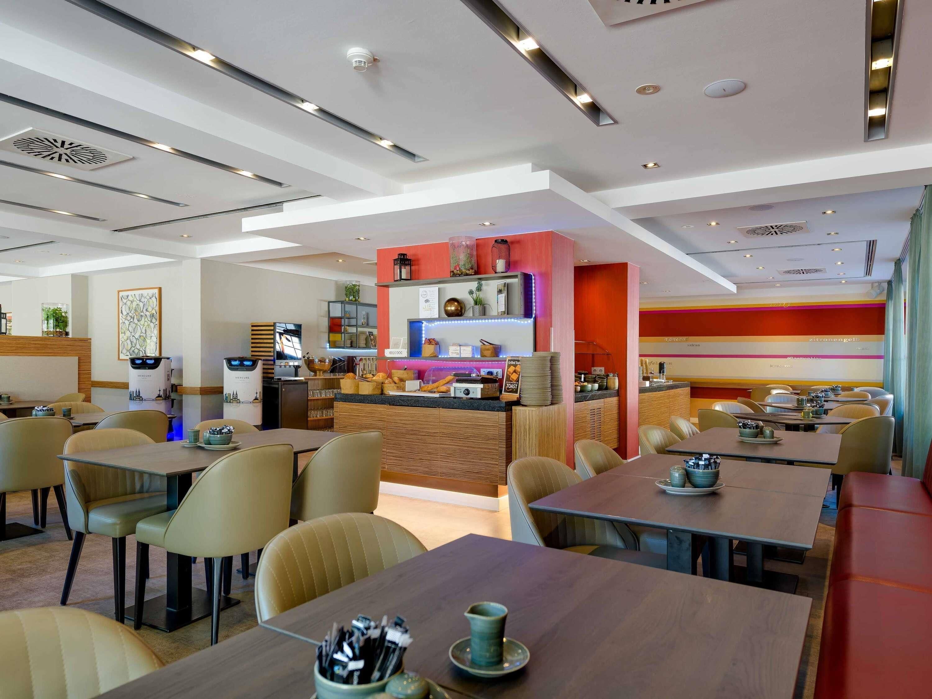 A modern restaurant with multiple dining tables and chairs, a bar area, and a kitchen in the background.