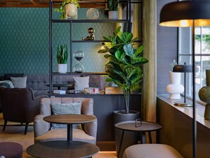 A modern living room with a sofa, two round tables, a bookshelf with plants, and a window with a lamp. - Mercure Hotel Köln Belfortstraße (Cologne)