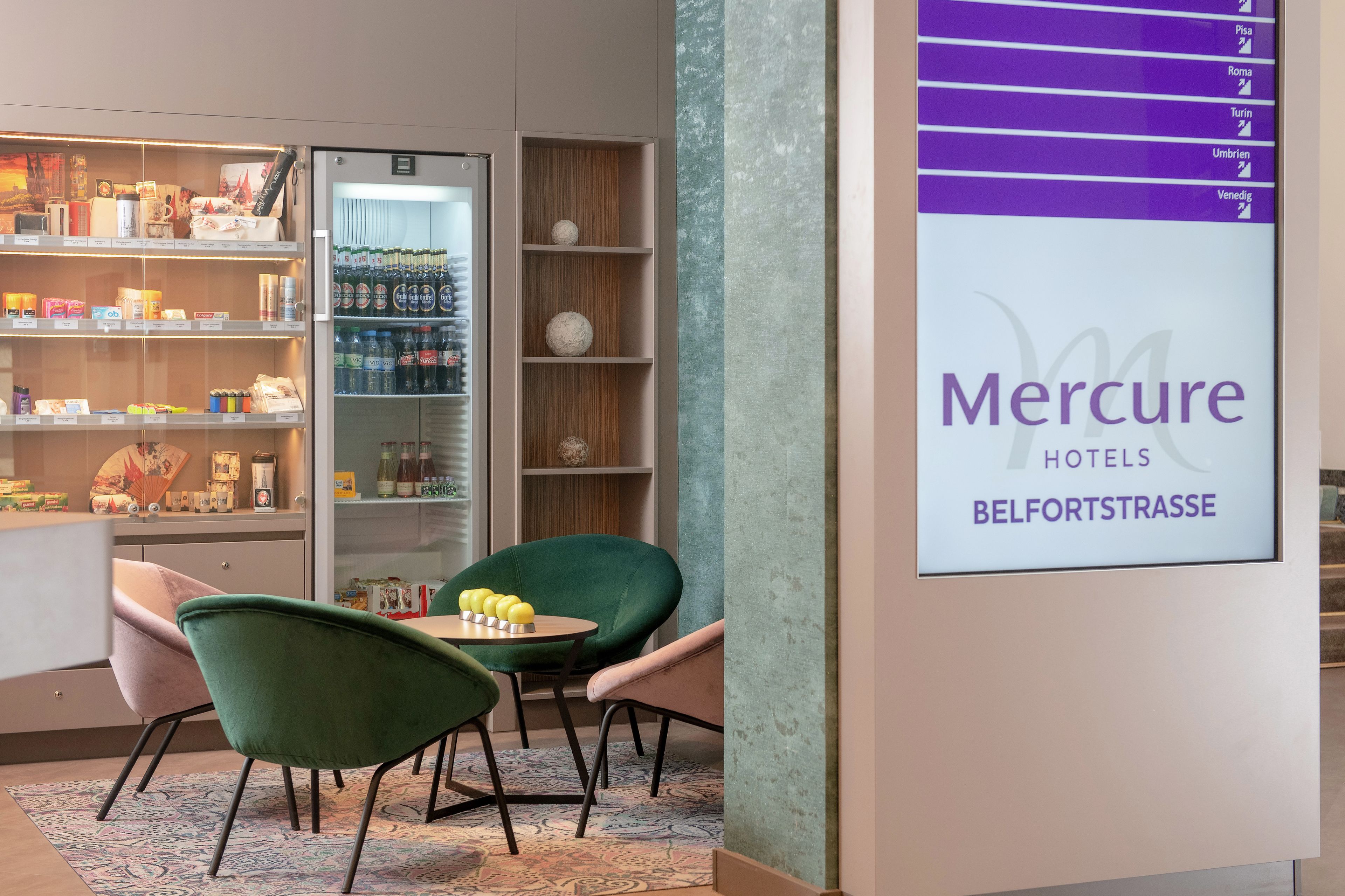 Un hall de hotel con un refrigerador de bebidas, una zona de estar con sillas verdes y una pared con un menú.