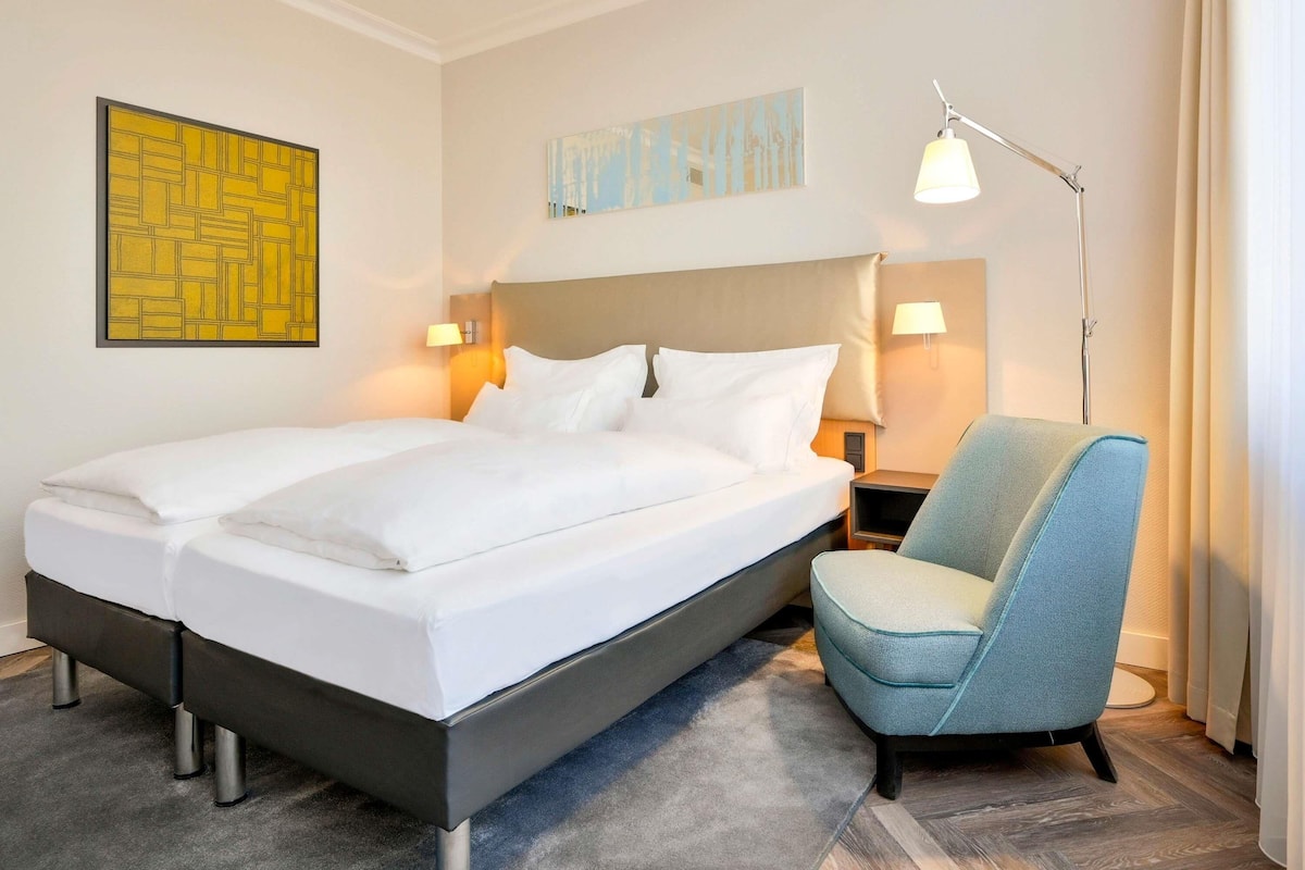 Una habitación de hotel con una cama, una silla, una mesita de noche, una lámpara y dos lámparas de pared.