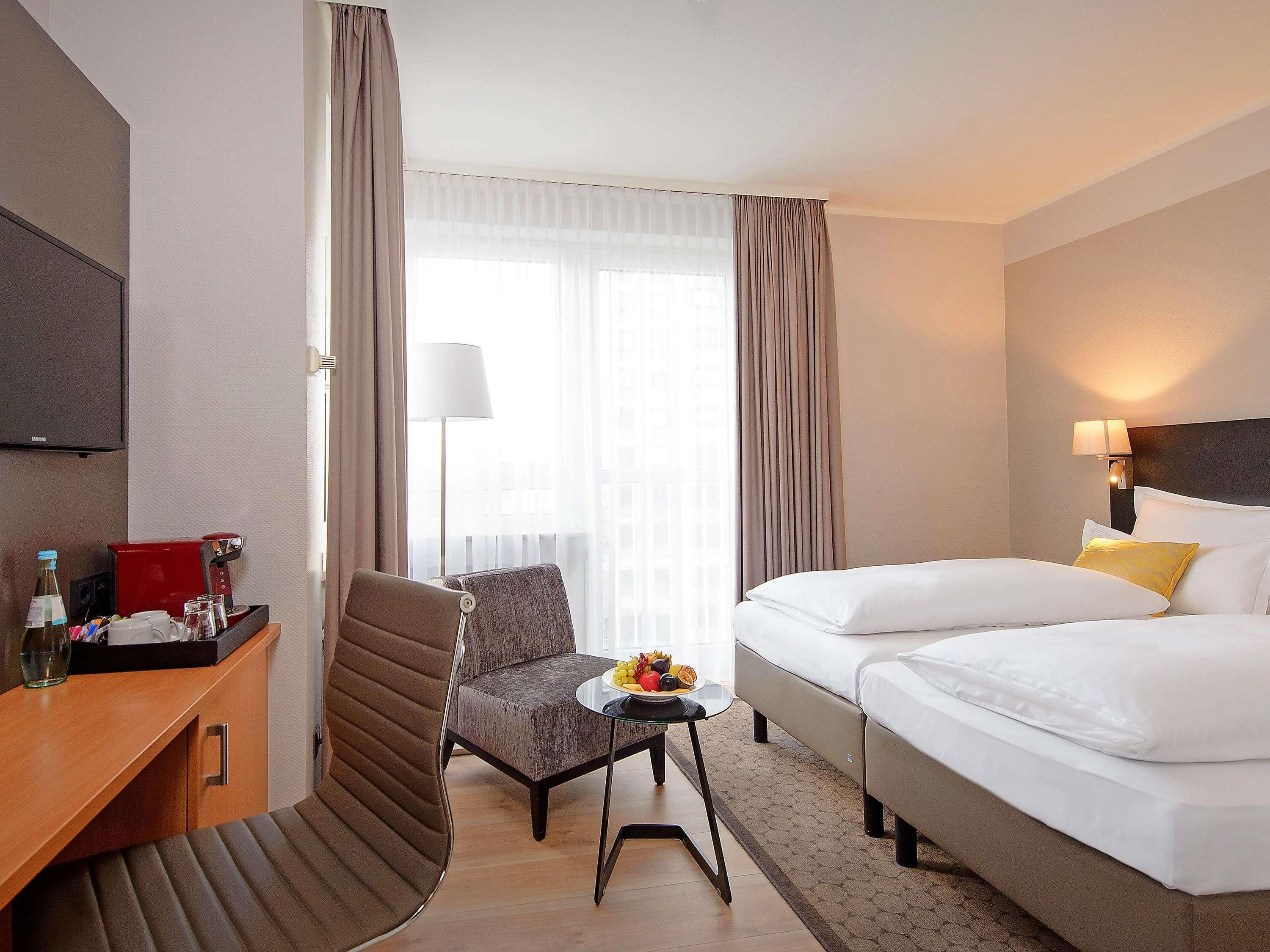 A hotel room with a bed, a desk with a coffee machine, a chair, and a small table with fruit.