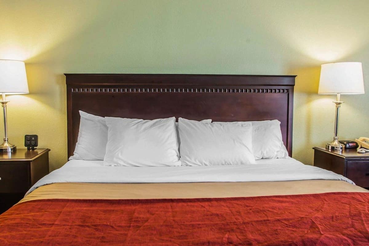 Una habitación de hotel con cabecera de madera, ropa de cama blanca, una manta roja y dos lámparas de noche.