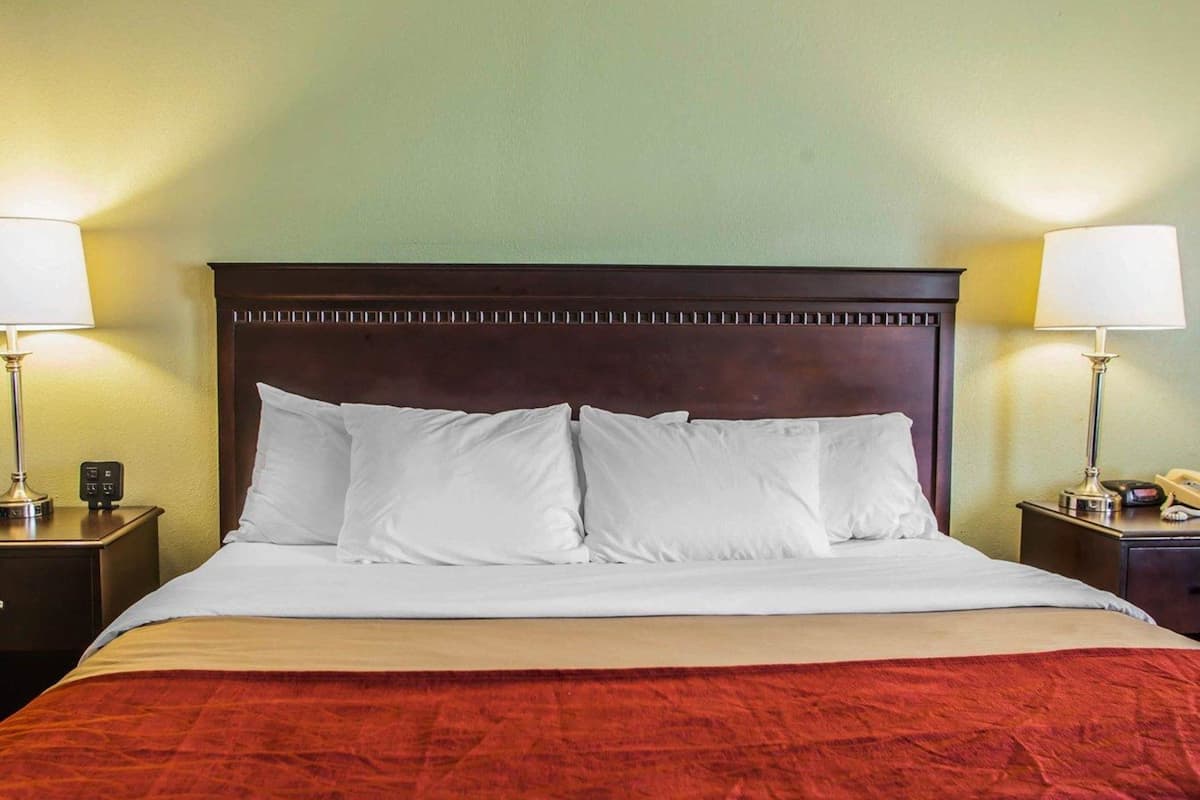 Una cama bien hecha con sábanas blancas y una colcha roja, flanqueada por dos mesitas de noche con lámparas y un reloj.