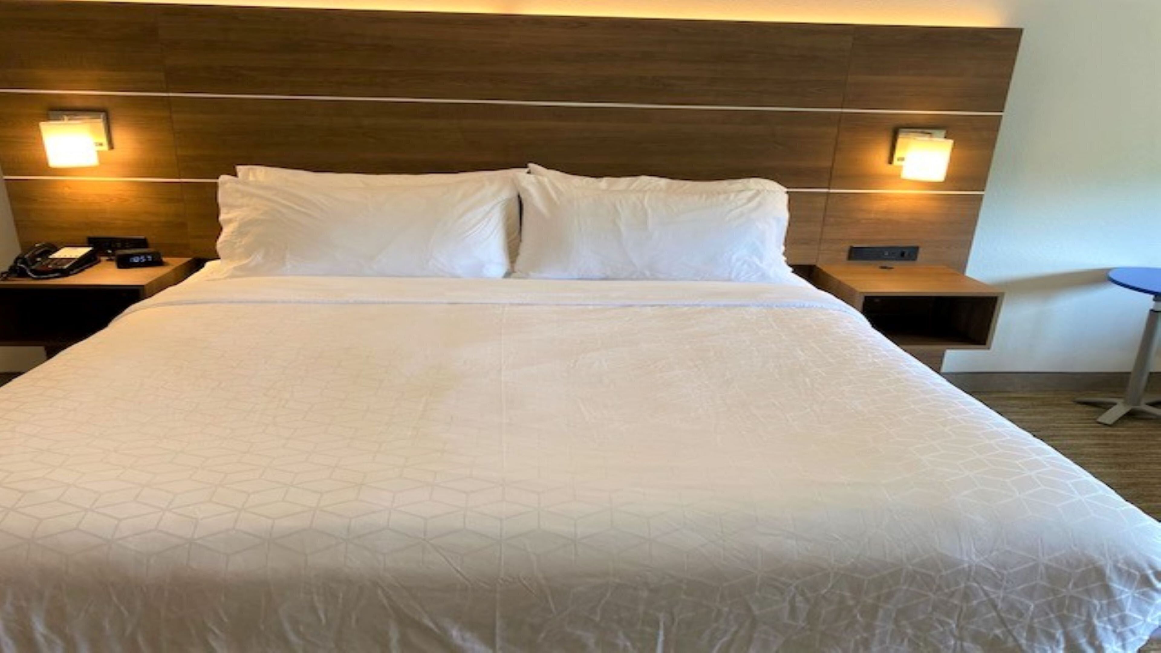 Un grand lit avec une tête de lit en bois, deux tables de chevet avec des lampes et une petite table ronde sur la droite.
