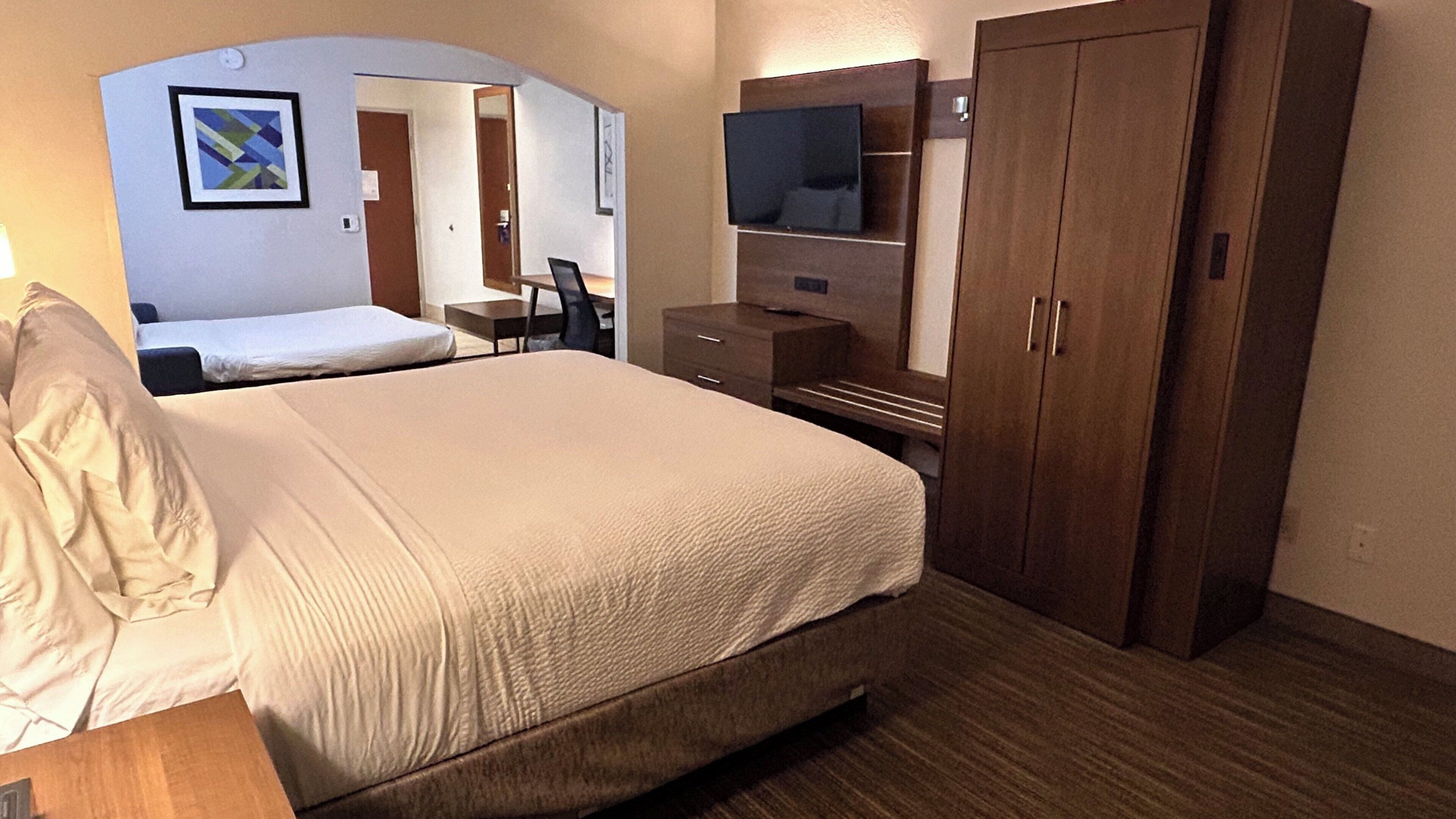 Habitación de hotel con una cama grande, un escritorio, una silla, un televisor y un armario.