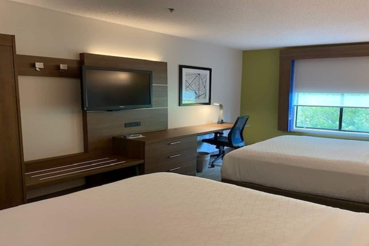 A hotel room with two beds, a desk, a chair, a television, and a window with a view of trees.