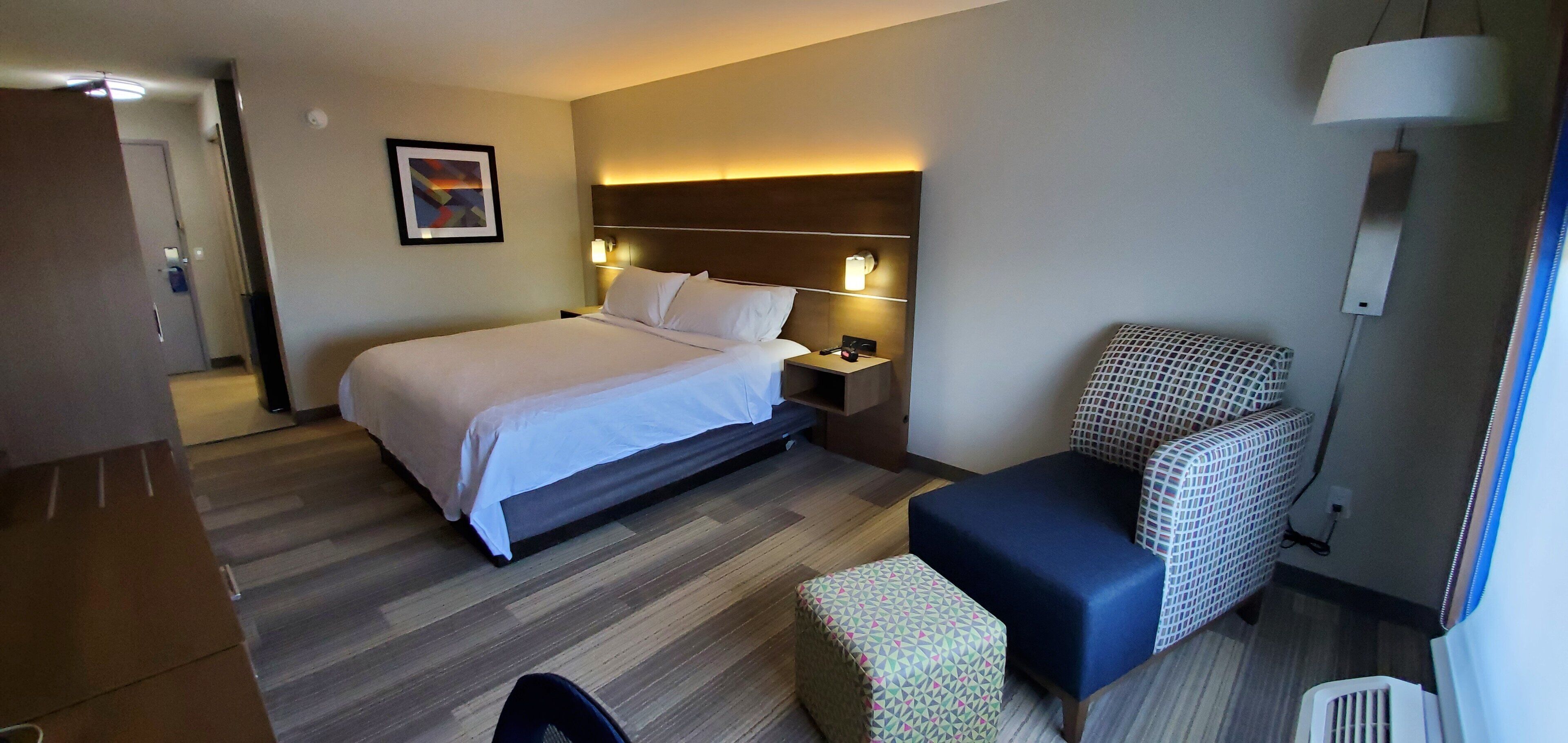 A hotel room with a bed, bedside table, lamp, and a small seating area with a patterned cushion.