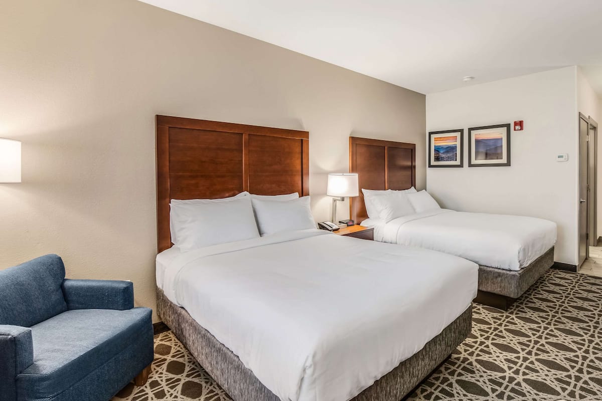 A hotel room with two beds, a blue armchair, a bedside table with a lamp, and a patterned floor.