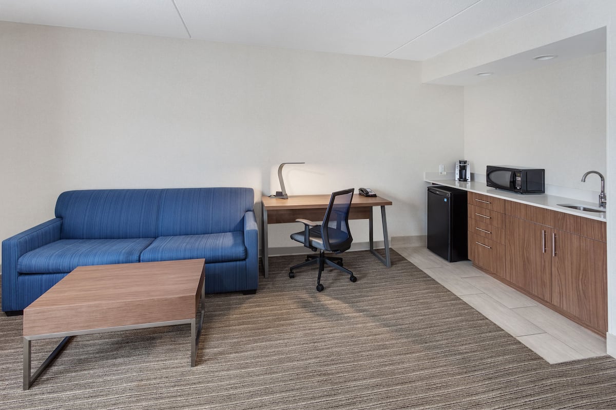 A compact living space with a blue sofa, a wooden coffee table, a desk with a chair, a microwave, and a small kitchenette.