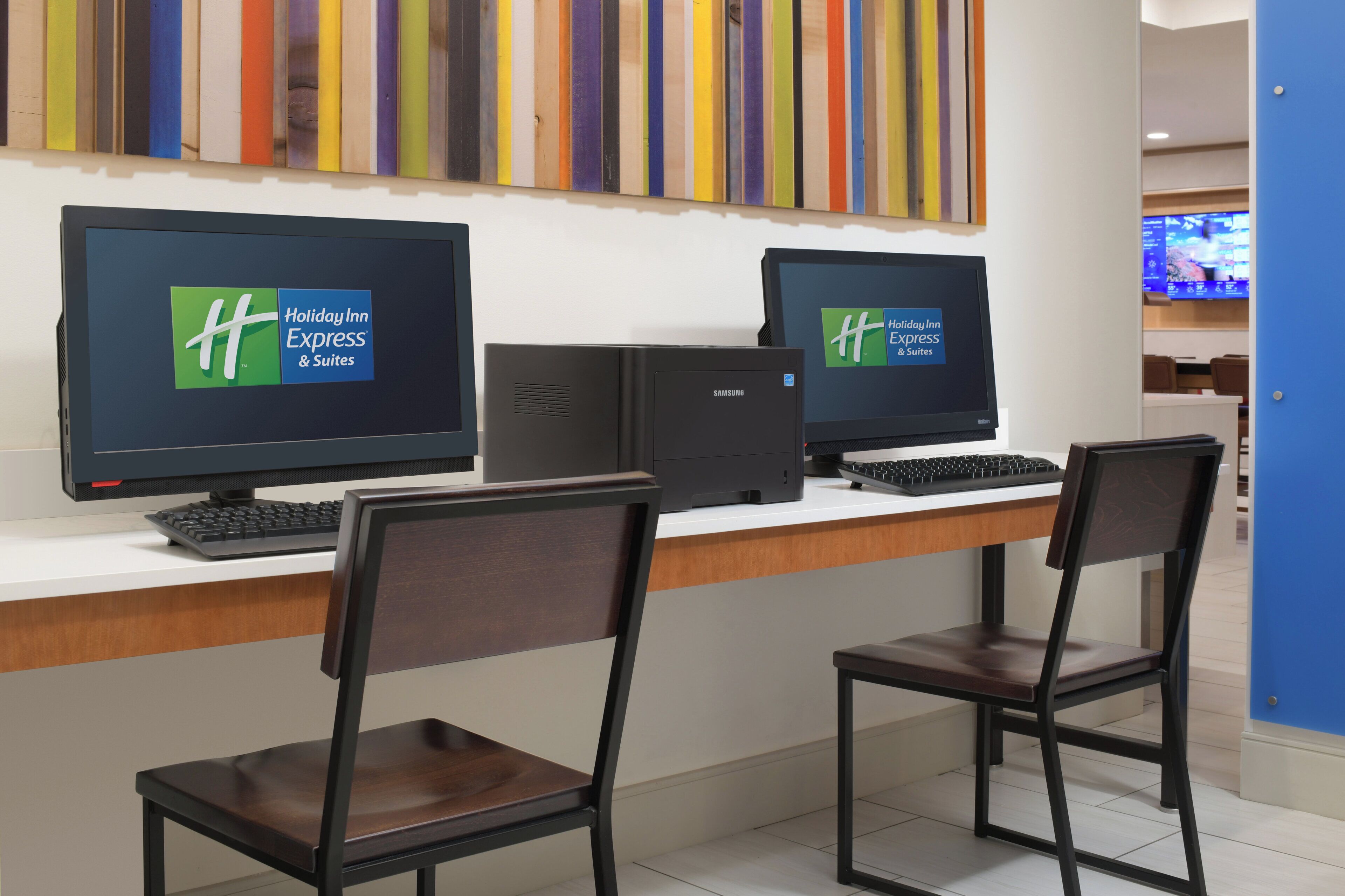 A hotel room with two monitors displaying the Holiday Inn Express & Suites logo, a desktop computer, and two wooden chairs.