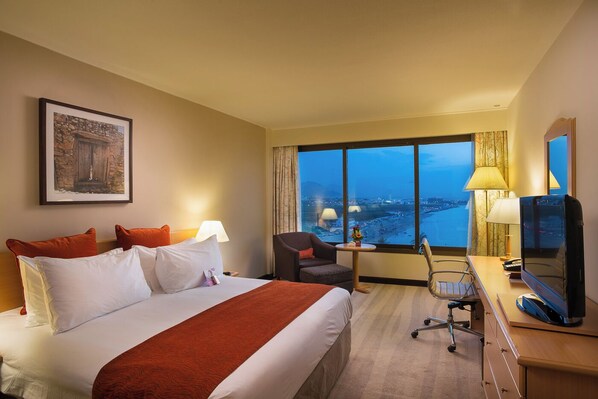 A hotel room with a large bed, a desk with a computer, a chair, and a view of a cityscape through large windows.