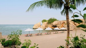 A beach with white umbrellas and lounge chairs, a palm tree, and rocky cliffs in the background.