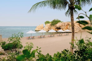A beach with white umbrellas and lounge chairs, a palm tree, and rocky cliffs in the background. - Crowne Plaza Muscat by IHG (Muscat)