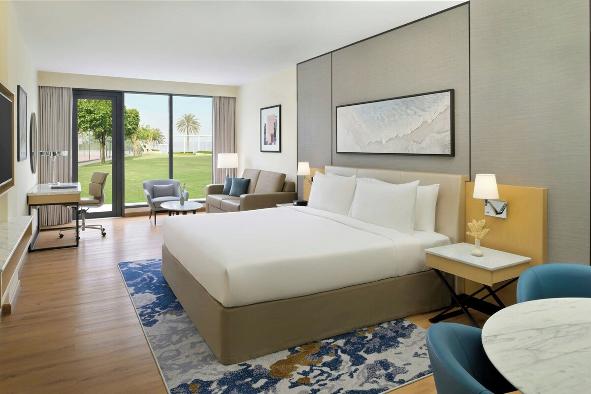 A hotel room with a large bed, a desk, a chair, a small table, and a view of the outdoors.
