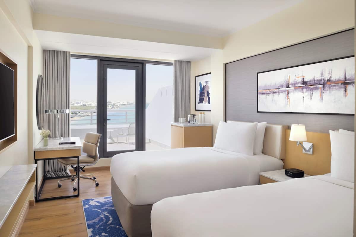 Ein Hotelzimmer mit zwei Betten, einem Schreibtisch, einem Stuhl, einem Fernseher, einem Fenster mit Blick auf die Stadt und einem Bild an der Wand.