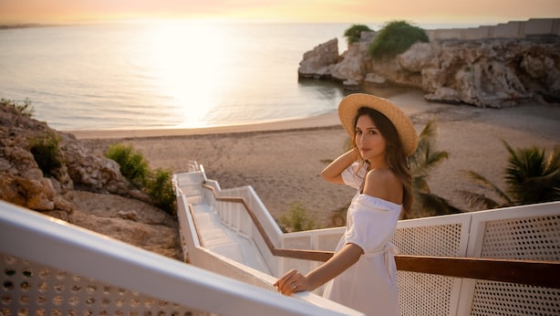 A person in a white dress standing on a balcony overlooking a beach with rocky cliffs and the sea.