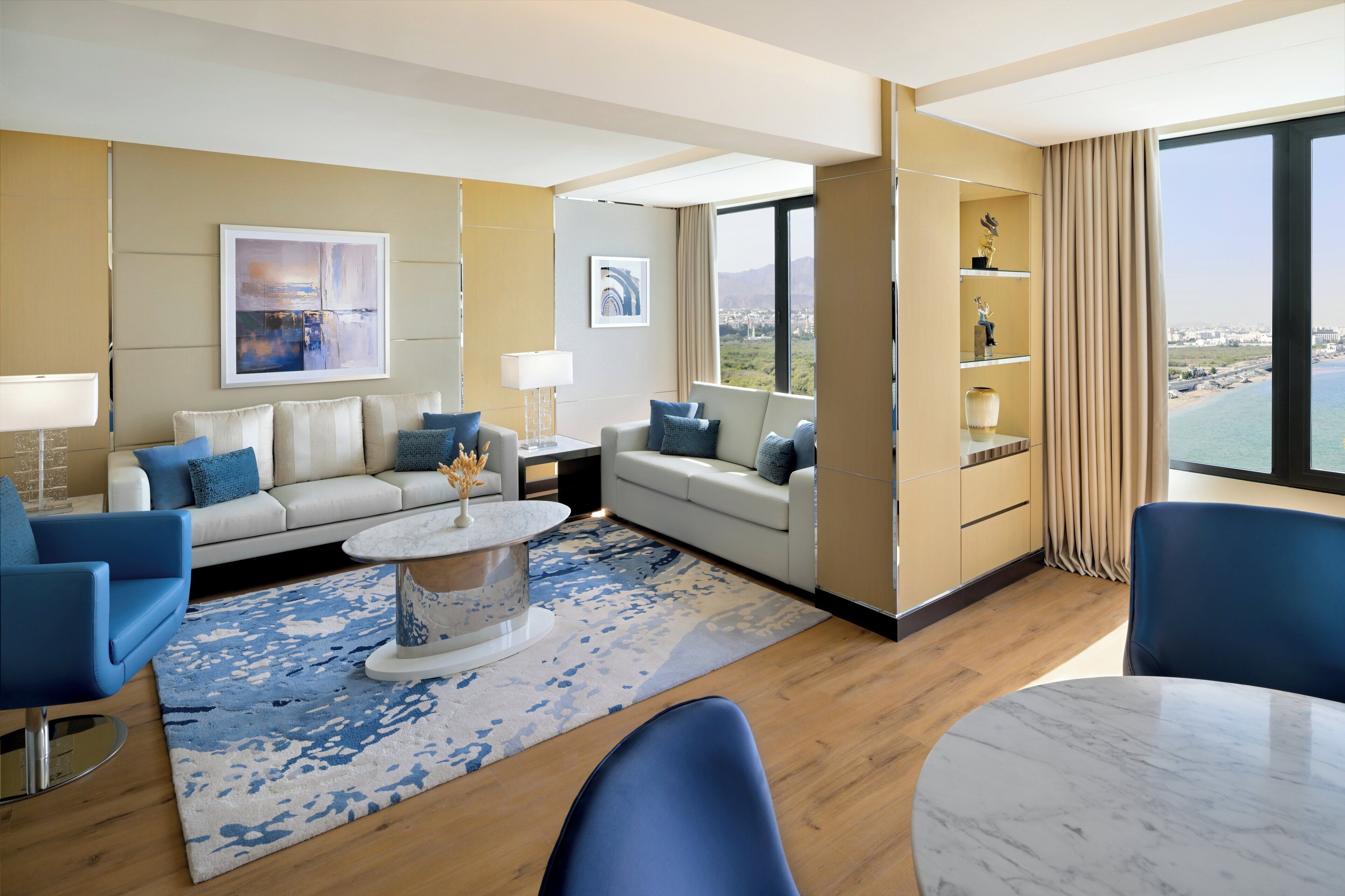 A modern hotel room with a large window offering a city view, a sofa set, a dining table, and a decorative shelf.
