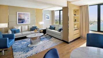 A modern hotel room with a large window offering a city view, a sofa set, a dining table, and a decorative shelf.