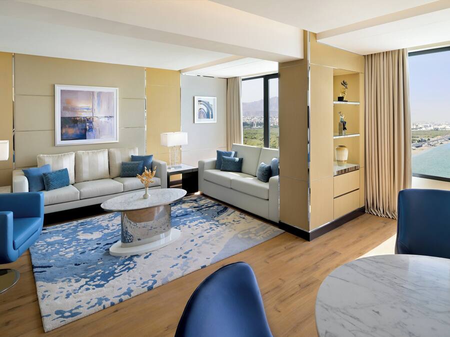 Una habitación de hotel moderna con un ventanal grande que ofrece vistas a la ciudad, un conjunto de sofás, una mesa de comedor y un estante decorativo.