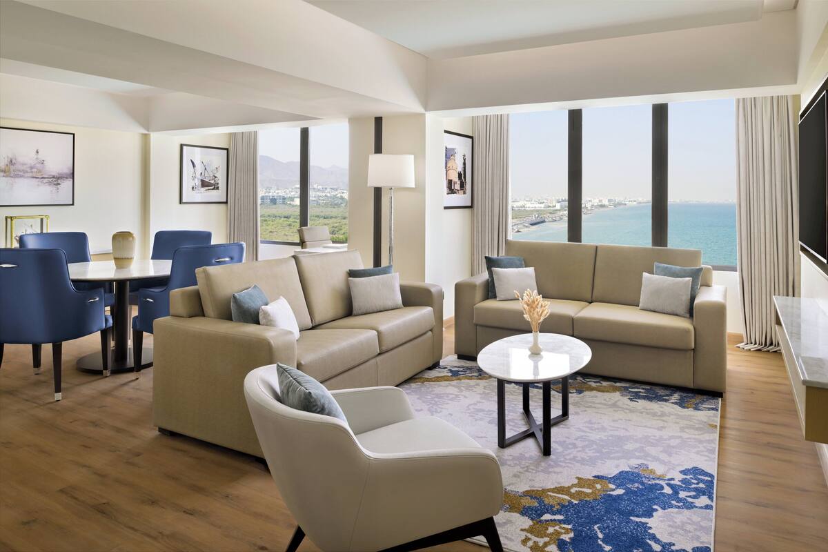 A modern hotel room with a large window offering a city view, a sofa, a chair, a small table, and a TV.