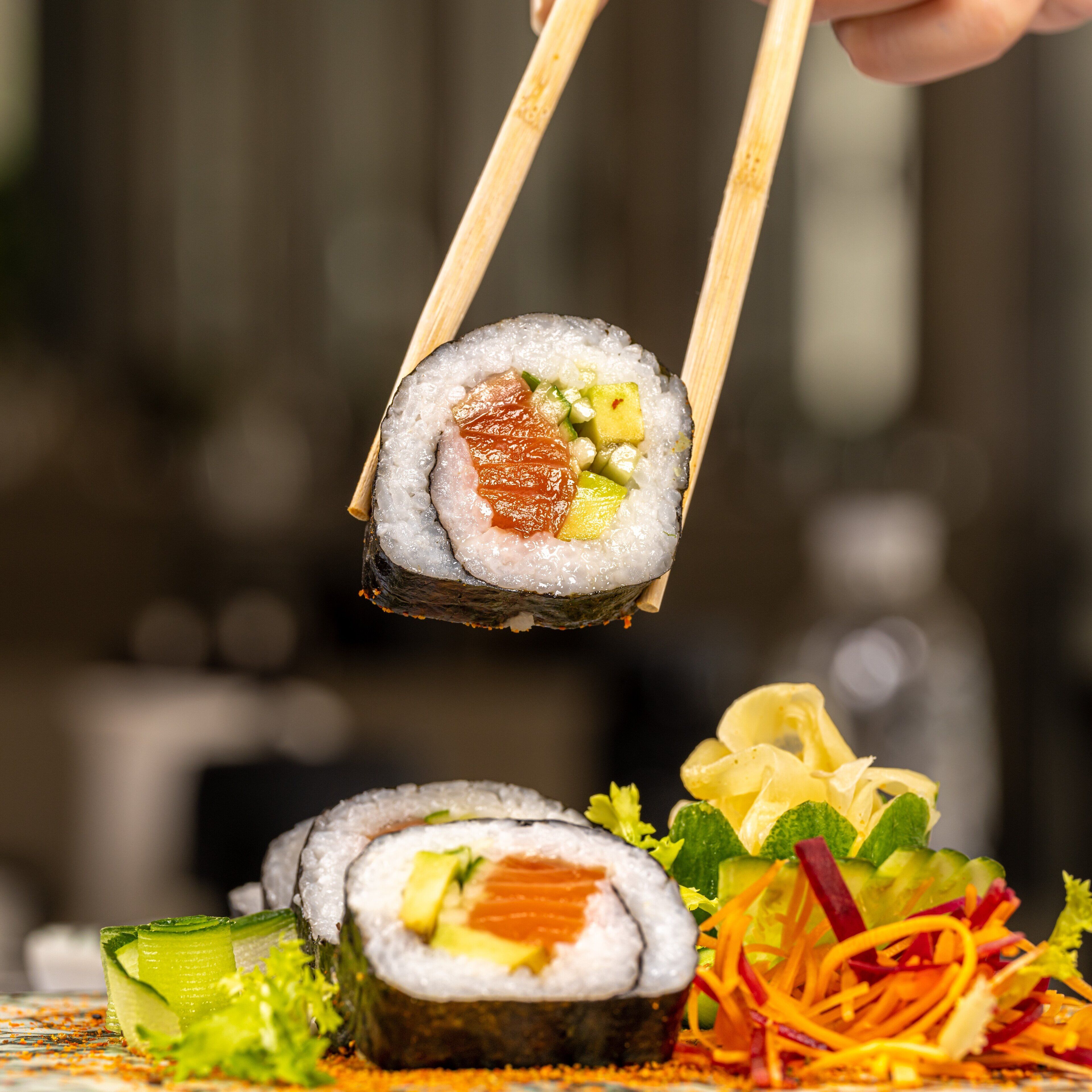 A sushi roll being held by chopsticks, with a side salad and cucumber slices on a plate.
